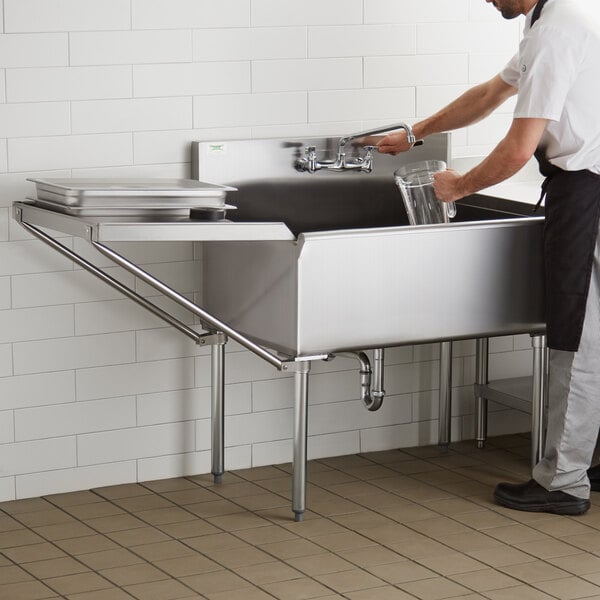 A man in a white shirt and black apron washing a glass in a stainless steel sink with a Regency detachable drainboard.