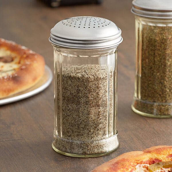 An American Metalcraft glass dredge with a stainless steel lid filled with spices on a table with a pizza.