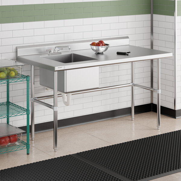 A stainless steel sink with an integrated work table, featuring a bowl of fruit and a utensil on the surface, set against a tiled wall in a commercial kitchen environment.