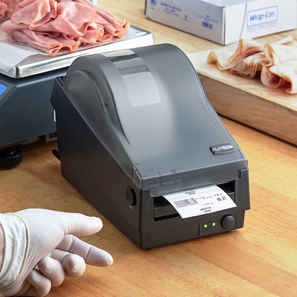 A black AvaTek thermal label printer printing a label, placed on a wooden counter near sliced deli meats.