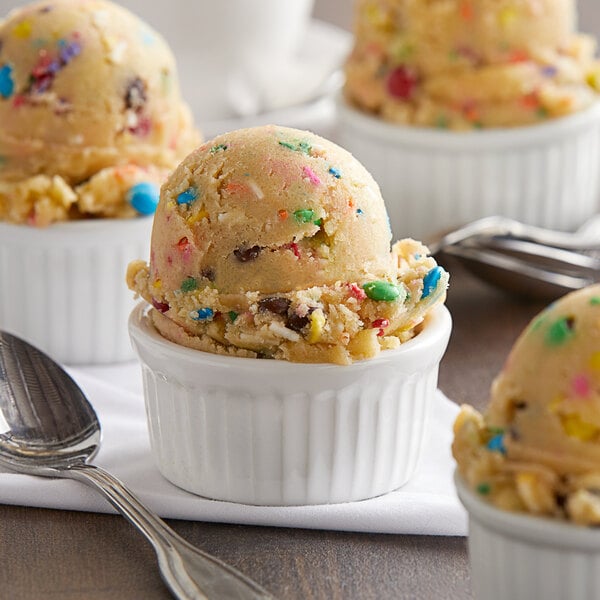 A white container of David's Cookies M&Ms Birthday Cake Edible Cookie Dough next to bowls of ice cream with colorful sprinkles and spoons.