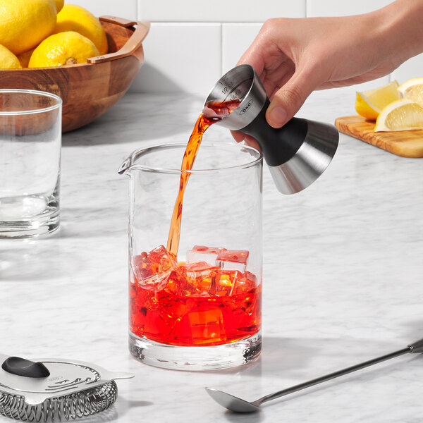 A hand using an OXO stainless steel double jigger to pour liquid into a glass.