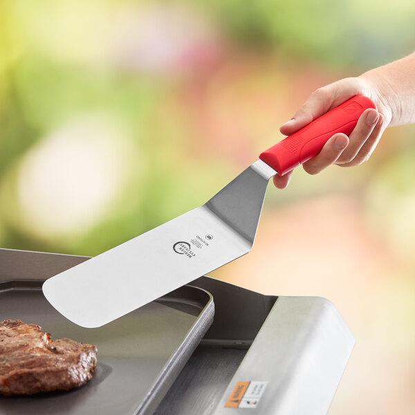 A hand holding a Mercer Culinary Millennia turner over a grill.
