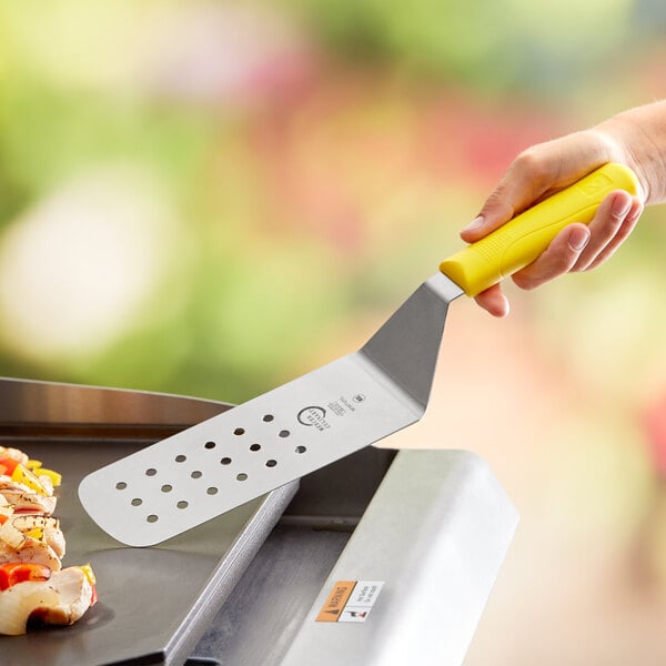 A hand holding a yellow Mercer Culinary turner over a grill.