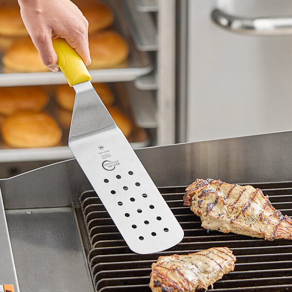 A person using a Mercer Culinary perforated turner with a yellow handle to cook chicken on a grill.