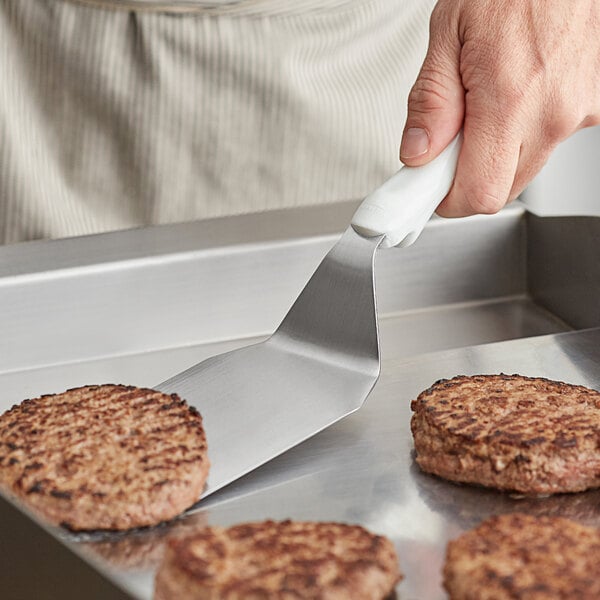 A person using a Vollrath stainless steel hamburger turner with a white handle to cook burgers on a pan.