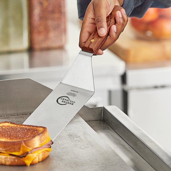 A person using a Mercer Culinary Praxis® turner with a rosewood handle to cut a grilled cheese sandwich.