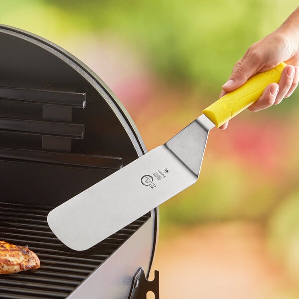 A hand holding a yellow Mercer Culinary Millennia turner over a grill.