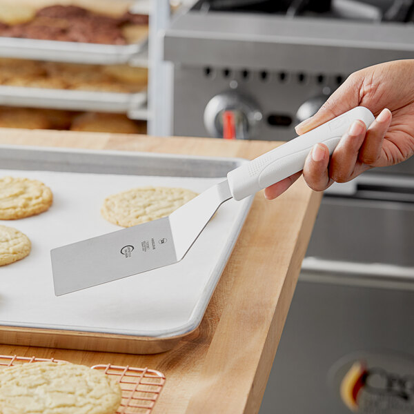 A hand holding a white Mercer Culinary square edge turner over cookies on a tray.