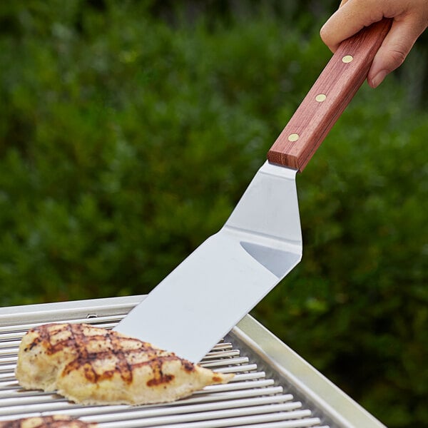 A hand holding a Dexter-Russell Traditional Solid Turner with a rosewood handle over a grill.