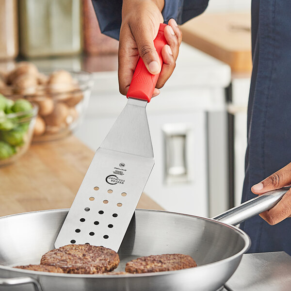 A person using a Mercer Culinary perforated spatula to cook meat patties in a pan.