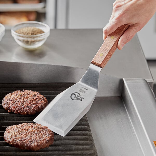 A hand holding a Mercer Culinary Praxis turner with a rosewood handle over a grill cooking burgers.