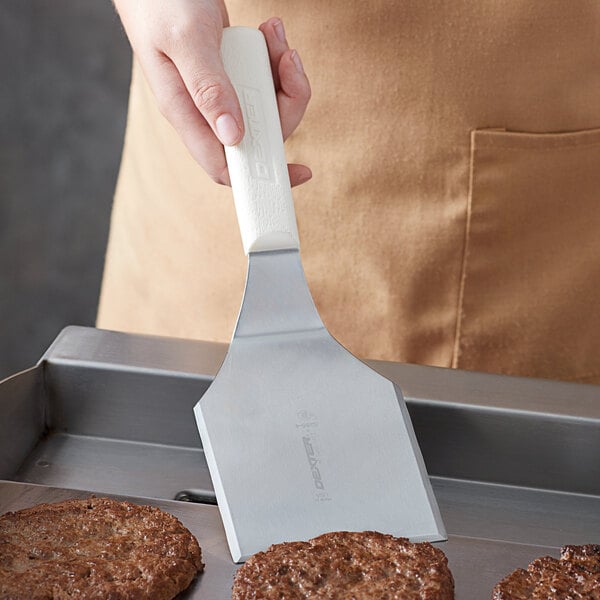 A person using a Dexter-Russell solid hamburger turner to cook burgers on a grill.