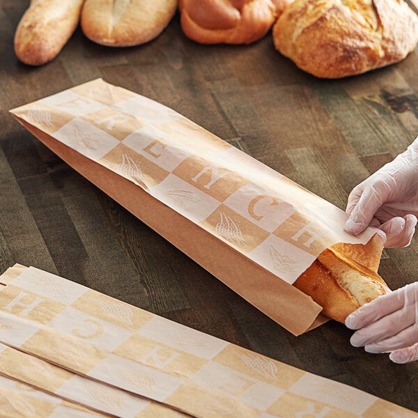 A hand in a glove putting a French loaf in a Bagcraft paper bag.