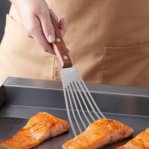 A person with a Dexter-Russell Rosewood Slotted Fish / Egg Turner holding a piece of cooked salmon over a pan.