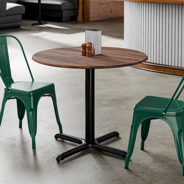A round standard height table with a textured walnut finish top and a black cross base plate, accompanied by green metal chairs.