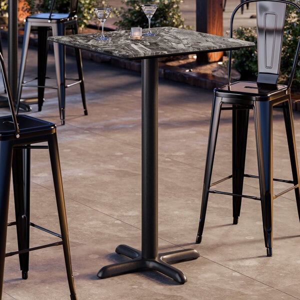 A square bar height table with a smooth, dark stone-like finish and a black cross base plate.
