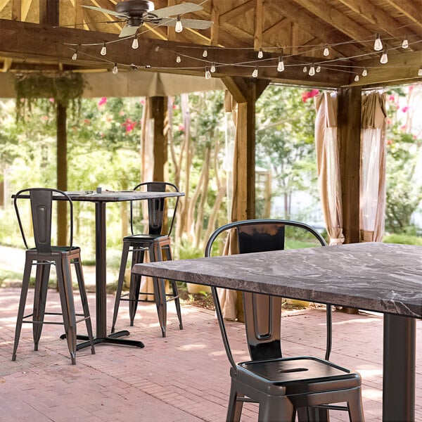 A Lancaster Table & Seating Excalibur bar height table with a smooth finish and cross base plate on a covered patio with chairs.