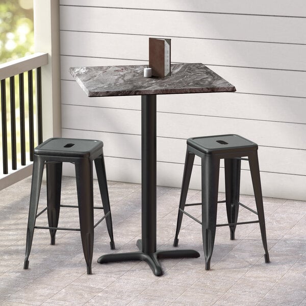 A Lancaster Table & Seating black metal square counter height table with a cross base plate and two black stools on a porch.