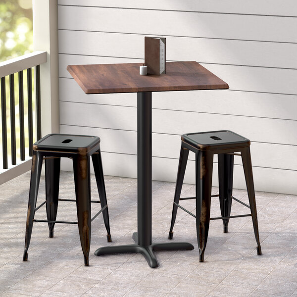 A Lancaster Table & Seating square counter height table with a textured walnut finish and cross base plate with two black stools on an outdoor patio.