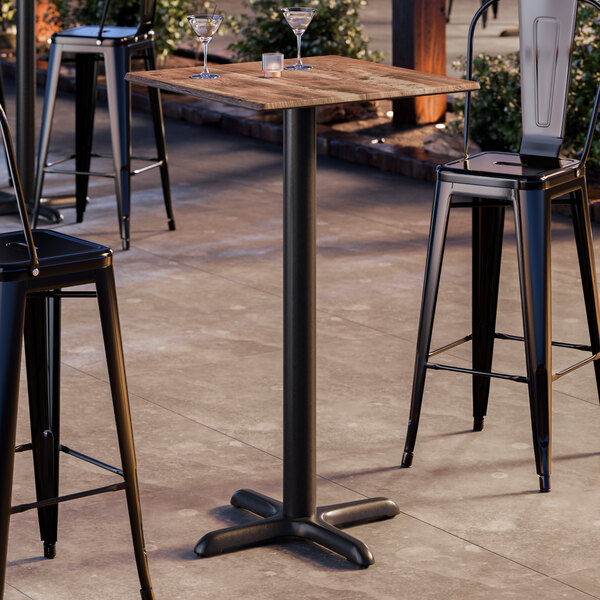 A square bar height table with a textured oak finish top and a black cross base plate.