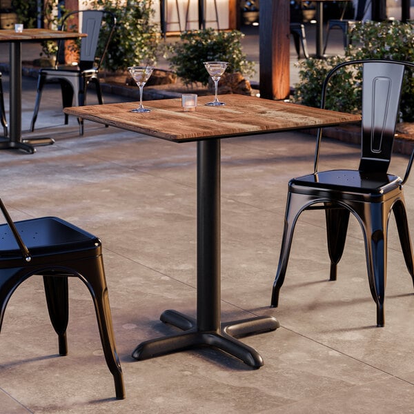 A square standard height table with a textured Yukon oak finish and a black cross base plate, shown with black metal chairs in an outdoor setting.