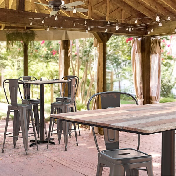 A Lancaster Table & Seating Excalibur bar height table with a textured mixed plank finish and cross base plate on a covered patio with chairs.