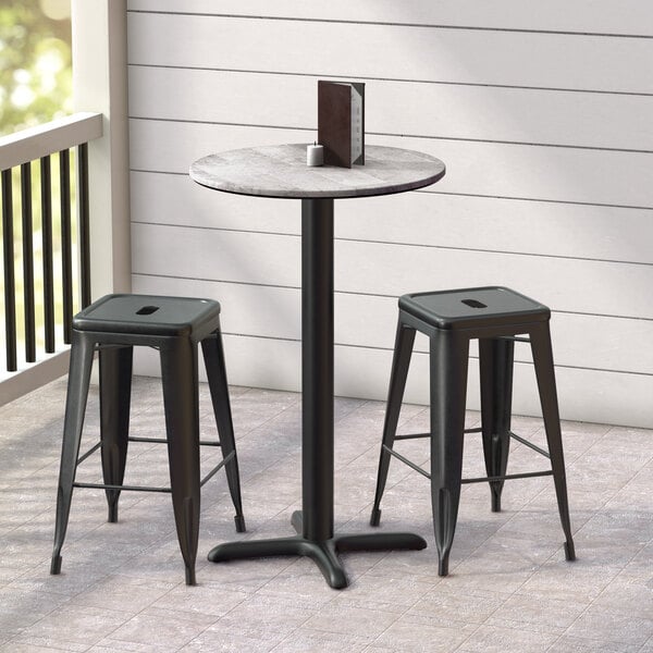 A Lancaster Table & Seating round outdoor table with a cross base plate and two black bar stools.