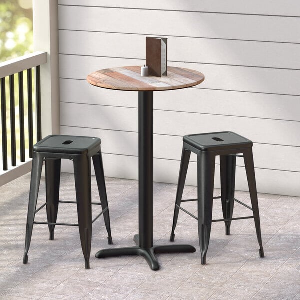 A Lancaster Table and Seating counter height table with a textured mixed plank finish and cross base plate on a porch with black bar stools.