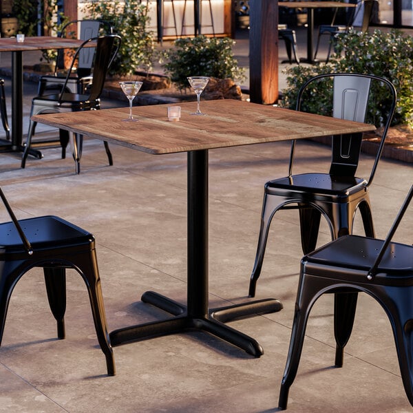 A square standard height table with a textured Yukon oak finish and a black cross base plate, surrounded by black metal chairs in an outdoor setting.