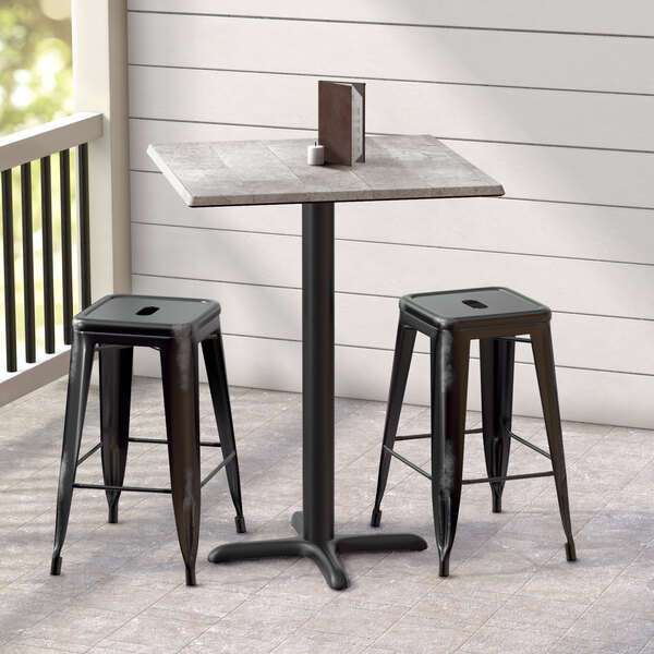 A Lancaster Table & Seating Excalibur square outdoor table with a textured finish and cross base plate with two black stools.