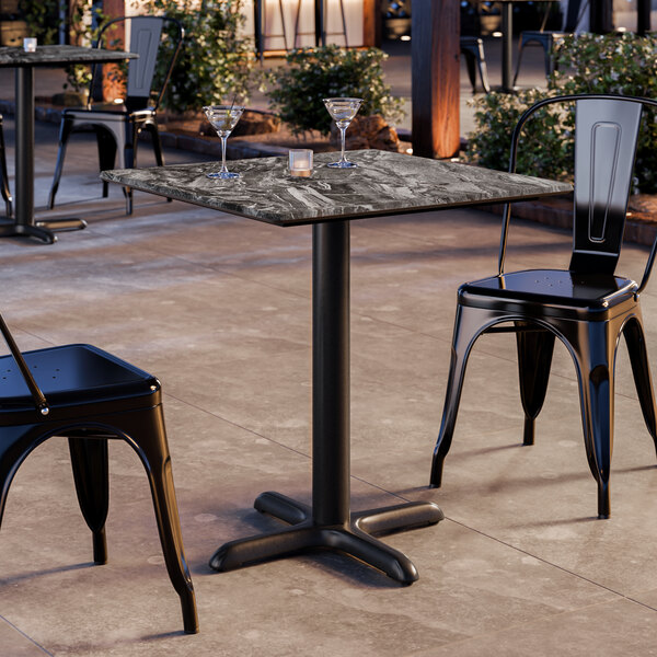 A square standard height table with a smooth Paladina finish and a cross base plate, set outdoors with two black metal chairs.