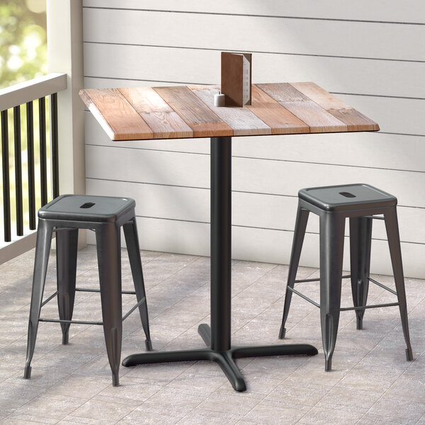 A Lancaster Table & Seating Excalibur counter height table with textured mixed plank finish and cross base plate, with two stools on a porch.