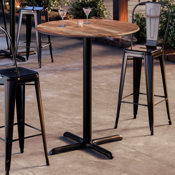 A round bar height table with a textured oak finish top and a black cross base plate.