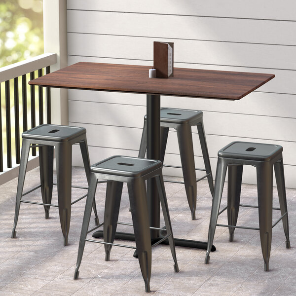 A Lancaster Table & Seating rectangular counter height table with a textured walnut finish and cross base plate surrounded by black stools on a tile floor.