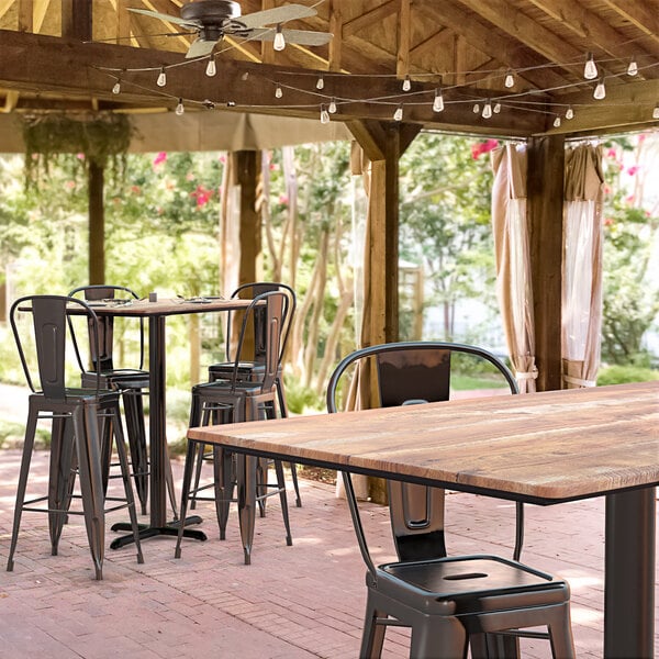 A Lancaster Table & Seating bar height table with a textured Yukon Oak finish and cross base plate on a brick patio with chairs.