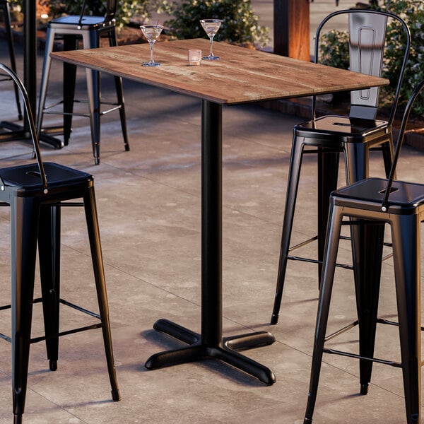A rectangular bar height table with a textured oak finish top and a black cross base plate.