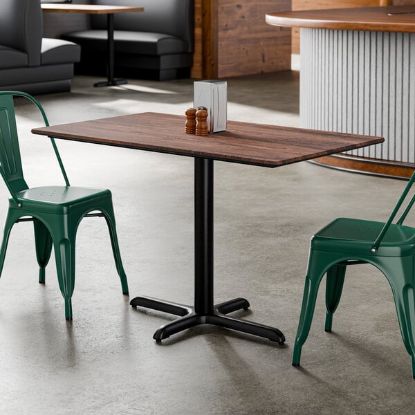 A rectangular standard height table with a textured walnut finish top and a black cross base plate, accompanied by green metal chairs.