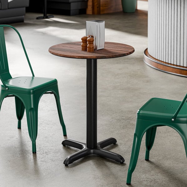 A round standard height table with a textured walnut finish top and a black cross base plate, accompanied by two green metal chairs.