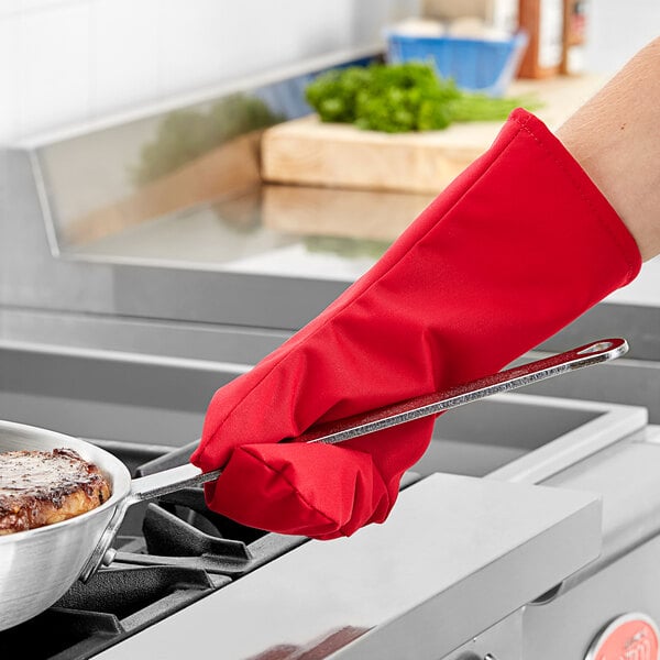 A person wearing a Tucker Safety red oven mitt holding a pan of food over a stove.