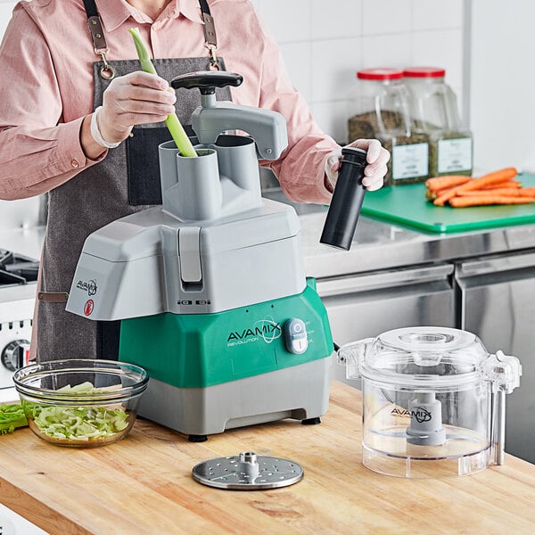 A woman using an AvaMix commercial food processor to slice green vegetables into a glass bowl.