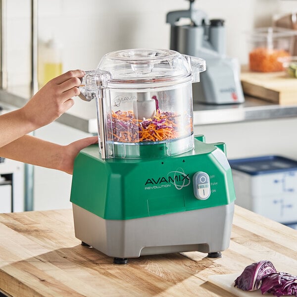 A green and gray AvaMix Revolution combination food processor with a clear plastic bowl, shown processing shredded vegetables on a kitchen counter.