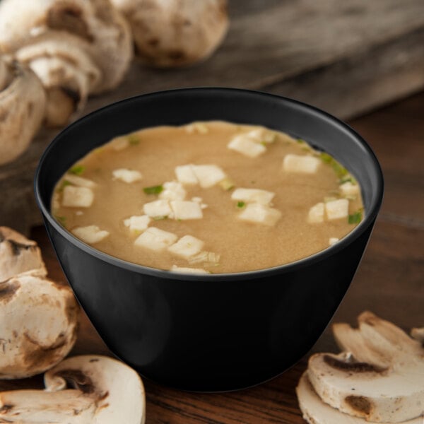 A Carlisle black melamine bouillon cup filled with soup and white cubes, next to mushrooms.