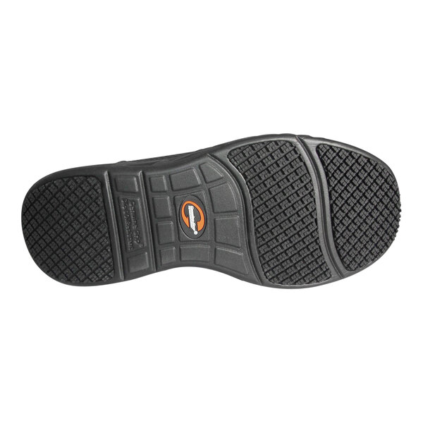 The image shows the black rubber outsole of a shoe with a textured, slip-resistant tread pattern and a small circular logo in the center.