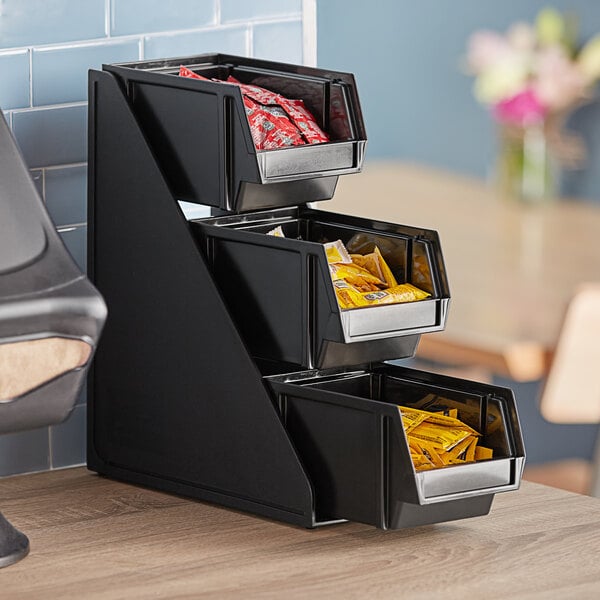 A black three-tier Vollrath condiment bin stand with black bins filled with condiments.