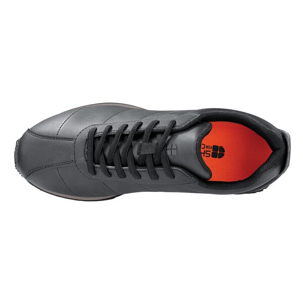 A black Shoe For Crews women's athletic shoe with orange laces and a white sole.