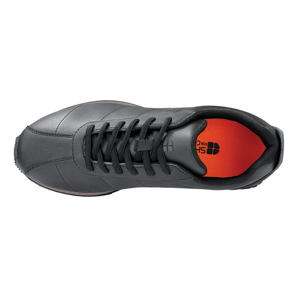 A black Shoe For Crews Avery sneaker with orange laces and a white sole.