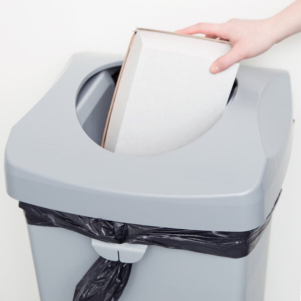 A hand putting a white box into a Rubbermaid Untouchable trash can.