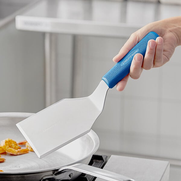 A hand holding a blue Dexter-Russell Sani-Safe hamburger turner over a pan.
