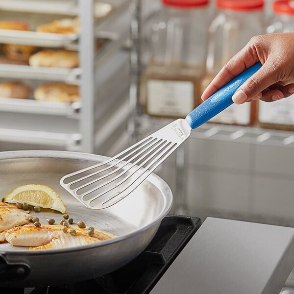 A hand holding a Dexter-Russell Sani-Safe blue slotted fish turner over a pan of food.